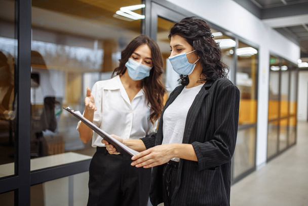 Due donne d'affari discutono di questioni di lavoro in ufficio indossando maschere mediche sterili. Lavora durante la quarantena pandemica di coronavirus. Nuovo concetto di misure normali, sanitarie e di sicurezza - Foto, immagini