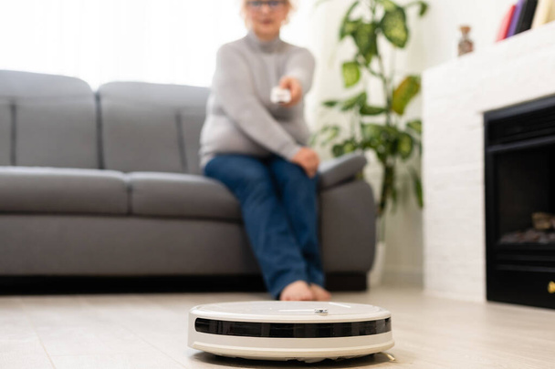 άτομα, αναψυχή, έννοια νοικοκυριού και τεχνολογίας - ηλικιωμένη γυναίκα, πάτωμα καθαρισμού με σκούπα ρομπότ στο σπίτι - Φωτογραφία, εικόνα