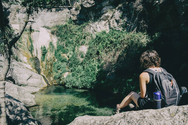 chica sentada en una roca mirando un paisaje de una pequeña cascada en un día soleado - Foto, imagen