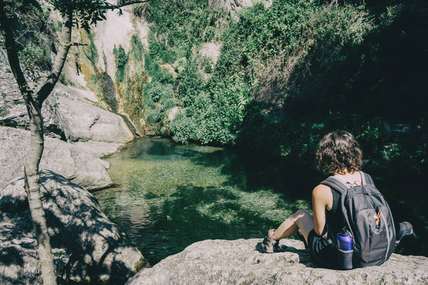 дівчина сидить на скелі, дивлячись на пейзаж маленького водоспаду в сонячний день
 - Фото, зображення