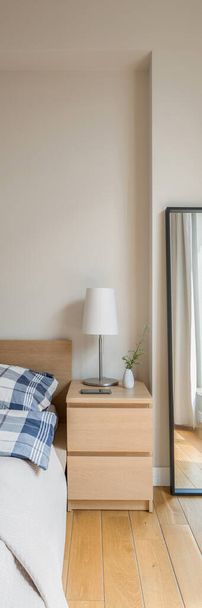 Vertical panorama of simple wooden bedside table with lamp next to bed and long mirror in black frame - Photo, Image