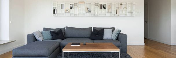 Panorama of decorative white shelf in living room with big gray, corner sofa, simple coffee table, fluffy carpet and nice wooden floor - Fotó, kép