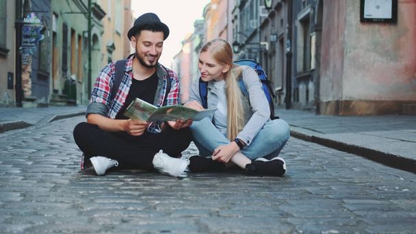 Young tourists couple using map, sitting on pavement and admiring historical surroundings - Photo, Image