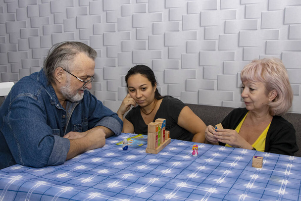 Kolmen hengen perhe pelaa lautapeliä pöydässä. Iäkkäät ihmiset kehittävät loogista ajattelua, eläkeläisten vapaa-aikaa, kodin viihdettä karanteeniaikana. - Valokuva, kuva