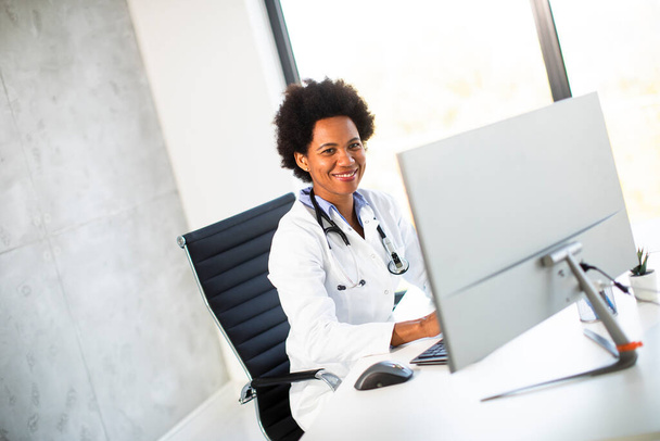 Female African American doctor wearing white coat with stethoscope sitting behind desk in the office - Φωτογραφία, εικόνα
