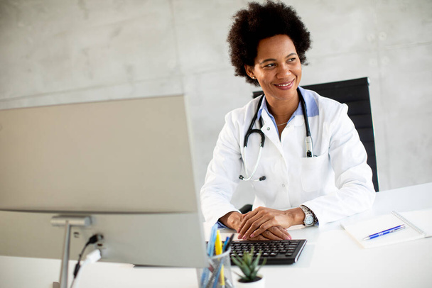 Nainen Afrikkalainen Amerikkalainen lääkäri yllään valkoinen takki stetoskooppi istuu pöydän takana toimistossa - Valokuva, kuva