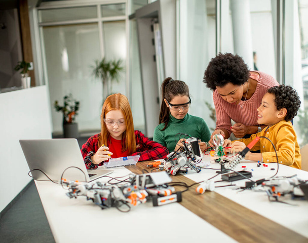 Grupo de niños felices con su profesora de ciencias afroamericana con computadoras portátiles programando juguetes eléctricos y robots en el aula de robótica - Foto, imagen