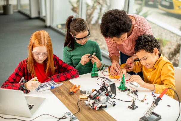 Группа счастливых детей со своей афроамериканской учительницей естественных наук с ноутбуками, программирующими электрические игрушки и роботов в классе робототехники - Фото, изображение