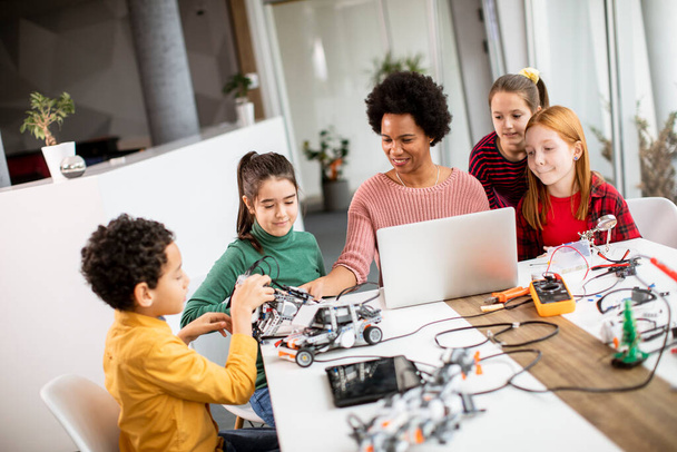 Группа счастливых детей со своей афроамериканской учительницей естественных наук с ноутбуками, программирующими электрические игрушки и роботов в классе робототехники - Фото, изображение