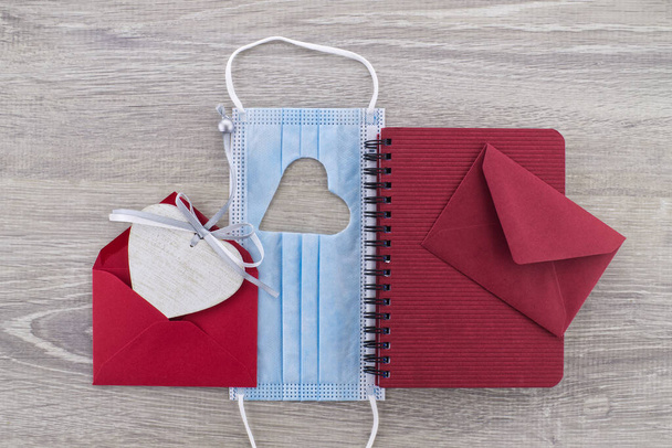 Face mask with cut out heart shape between a wire bound notepad and red envelope on wooden background with free copy space. Social distancing and contact-less greetings - Photo, Image