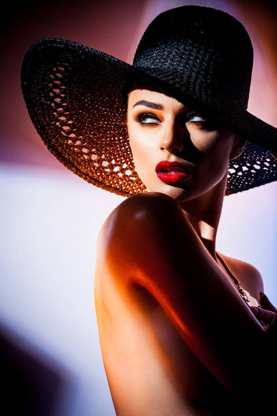 Fashion concept. Portrait of a beautiful woman in big black hat in bright mixed contrast light. Female model wearing accessories poses in studio. Professional makeup and colorful background. - Fotografie, Obrázek
