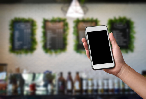 Teléfono móvil en blanco con pantalla táctil en la mano con fondo borroso de la cafetería, concepto para el uso de dispositivos digitales en la vida cotidiana, ordenar comida y bebida, en todo el mundo. - Foto, imagen