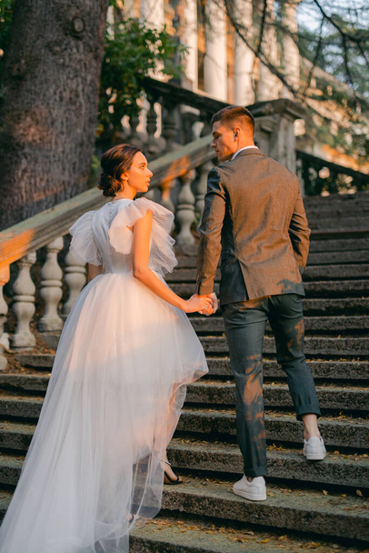 Full length portrait of handsome man in stylish suit embracing his charming young bride in white wedding dress. Happy newlywed couple posing against the background of a stone staircase in the park. - Foto, imagen