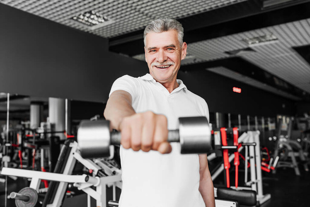 Sport. Close up in focus of a senior man who is smiling and extends his hand with a dumbbell to the camera. Smiling gray-haired man in the gym. - Foto, Bild