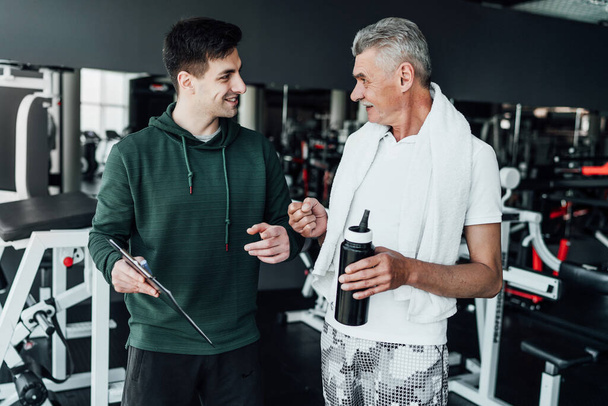 Two men in the gym are talking, one of them the coach shows the training plan to the older man. Sports Hall. Training plan. Coach. Rehabilitation. - Fotoğraf, Görsel