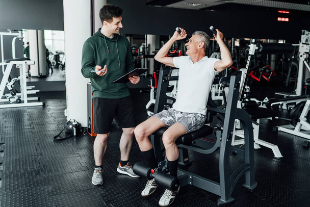 Photo in the gym, a senior man performing an exercise on a simulator, his rehabilitation coach observes and supports him, holding a training plan in his hand. - Foto, Imagen