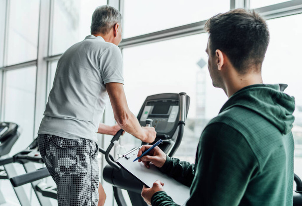 The focus is on a training plan held by a male coach. The photo shows two men in the gym, one of them performing an exercise on a simulator, the other watching. - Foto, Bild