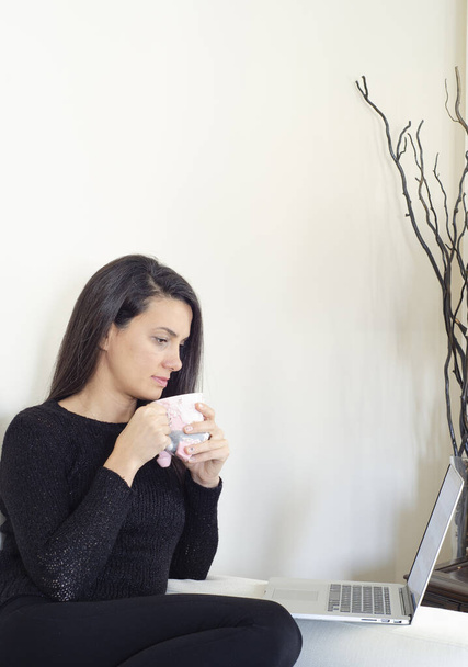 Krásná mladá žena pracující na notebooku, zatímco sedí v obývacím pokoji, pití kávy - Fotografie, Obrázek