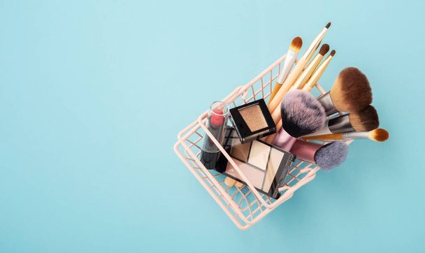 Makeup beauty products in pastel pink shopping basket on blue background, flat lay with copy space - Photo, Image