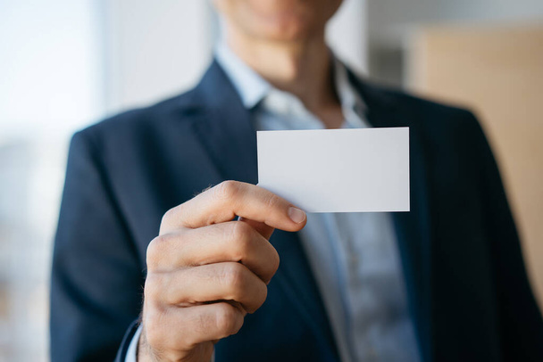 Hombre sosteniendo la tarjeta de visita en blanco con espacio para copias, de pie en la oficina moderna. Mock up, concepto de publicidad - Foto, imagen