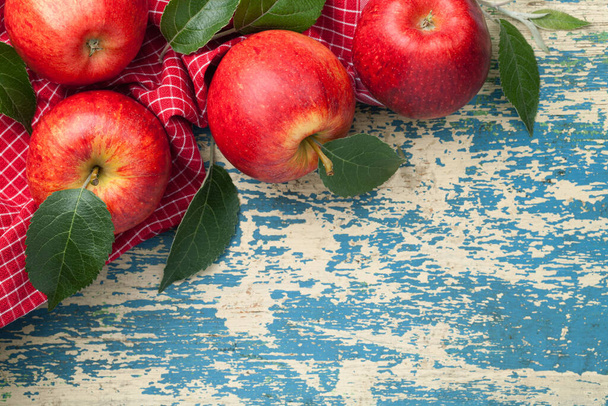 Red apples, fresh green leaves composition on red and white checkered tablecloth. Copy space on wooden background. View from above - Фото, изображение