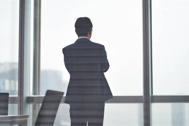 traseiro vista de um asiático homem de negócios de pé na frente de escritório janela pensando braços cruzados - Foto, Imagem