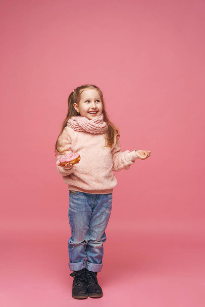 little girl in a fluffy sweater and a headband, smiling with a doughnut in her hand. Sweet appetizer, delicious pastries. a child shows a delicious dessert on a pink background, full-length in studio - Foto, immagini