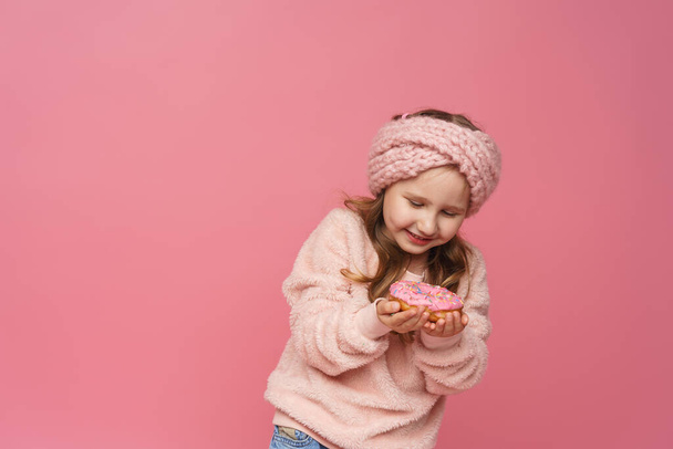 little girl in a fluffy sweater and a headband, smiling with a doughnut in her hand. Sweet appetizer, delicious pastries. a child shows a delicious dessert on a pink background in studio - Photo, image