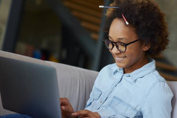 Erfolgreicher Schüler. Fröhliches Teenie-Schulmädchen mit Brille und Bleistift im Haar, lächelnd, während sie Laptop zum Lernen benutzt, zu Hause auf der Couch sitzend - Foto, Bild