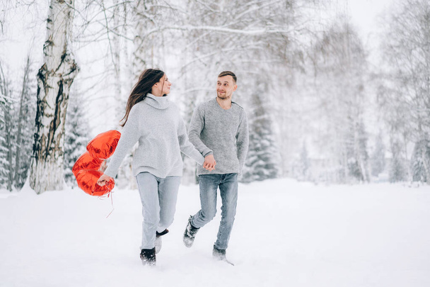 Una pareja amorosa corre a través de la nieve en el bosque. Mujer sosteniendo globos en forma de corazón en sus manos. - Foto, Imagen