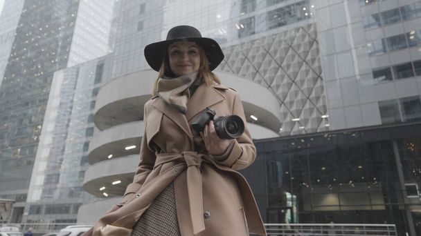 Привлекательная молодая блондинка с камерой в руках на городской улице. Начали. Элегантная женщина в коричневом пальто и черной шляпе улыбается и позирует в зимний день на фоне современных зданий - Фото, изображение