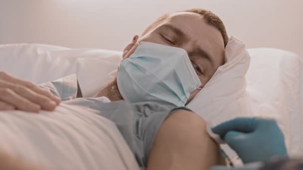 Primer plano de un paciente caucásico joven con máscara facial acostado en la cama, mirando a la cámara, luego a mano con un guante azul de un trabajador médico irreconocible que le da una dolorosa inyección de vacuna  - Metraje, vídeo