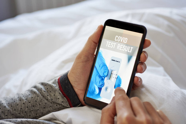 detailní záběr mladého bělocha v pyžamu, ležícího v posteli, kontrolujícího výsledek svého covidového testu v simulované aplikaci v jeho smartphonu, který čte výsledek kovidního testu - Fotografie, Obrázek