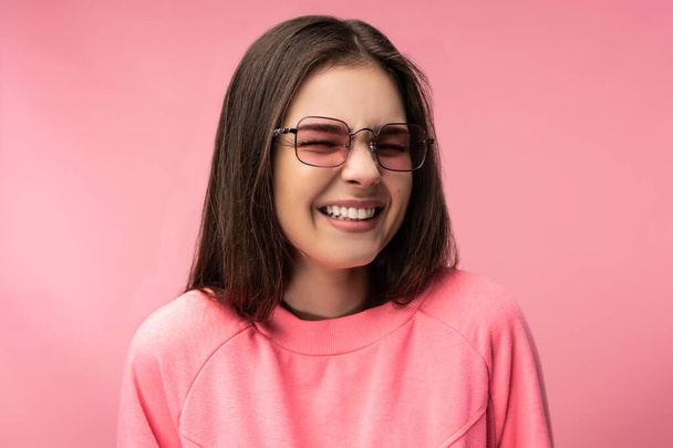 Photo of attractive young woman in glasses laughs and smiles, wears casual pink t-shirt white pants isolated pink color background - Φωτογραφία, εικόνα