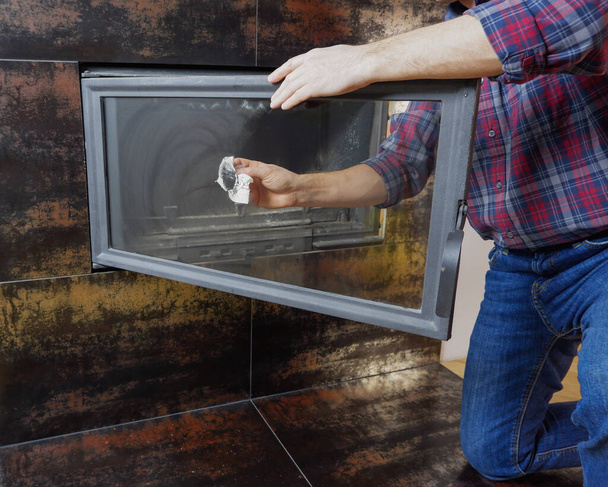 A man in a shirt and blue jeans cleans the heat-resistant glass of a modern fireplace. Close-up view of the hands. - Photo, Image