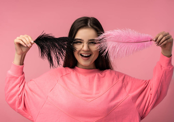 Foto de mujer joven atractiva en gafas cubre la cara por plumas y sonrisas. Lleva casual camiseta rosa pantalones blancos aislados color rosa de fondo - Foto, imagen