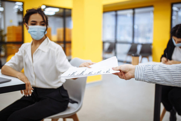 Young colleagues pass documents to each other working at an office during coronavuris pandemic outbreak. Professional workers wearing medical masks checking news and trends in business - Foto, afbeelding