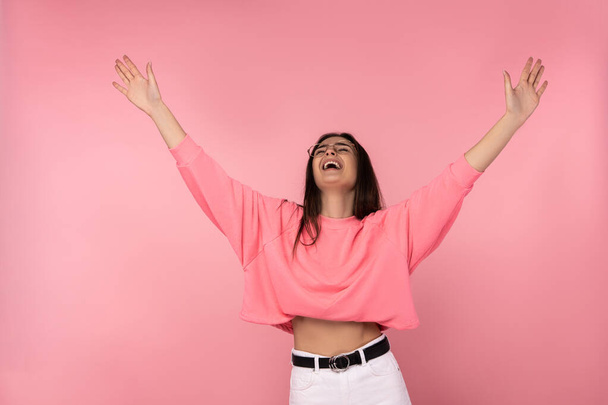 Foto de mulher atraente em saltos óculos com as mãos para cima, usa casual rosa t-shirt calças brancas isolado cor de fundo rosa - Foto, Imagem
