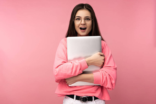 Kuva houkutteleva nainen lasit, yllättynyt suu auki, pitää kannettava tietokone ja pukeutuu rento vaaleanpunainen t-paita valkoiset housut eristetty vaaleanpunainen tausta - Valokuva, kuva