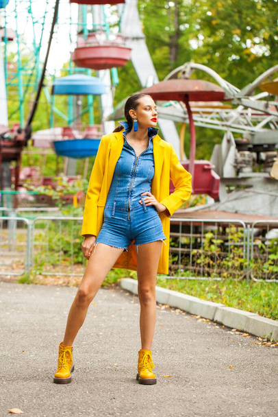 Fashion model in yellow coat and denim overalls posing on carousel background in an amusement park - Fotoğraf, Görsel