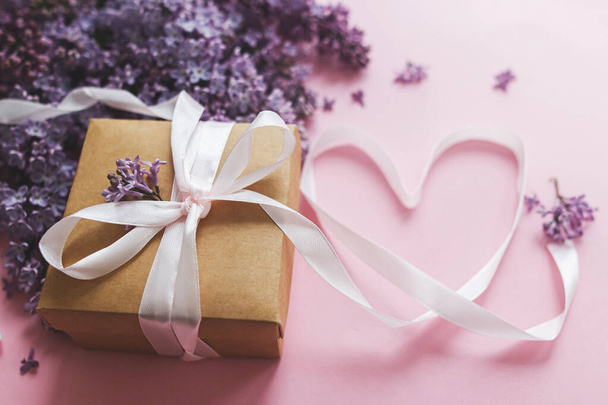 Lilac flowers, gift box and heart ribbon on pink paper. Stylish floral greeting card. Happy valentines day and mothers day concept. Purple lilac bouquet with stylish craft present - Photo, Image