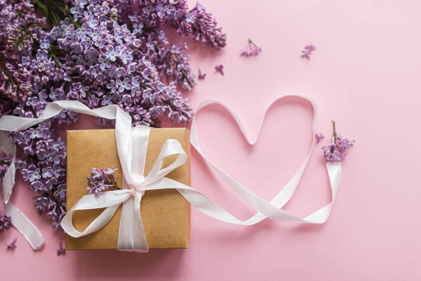 Lilac flowers, gift box and heart ribbon on pink paper, flat lay. Happy valentines day and mothers day concept. Purple lilac bouquet and craft present - Photo, Image