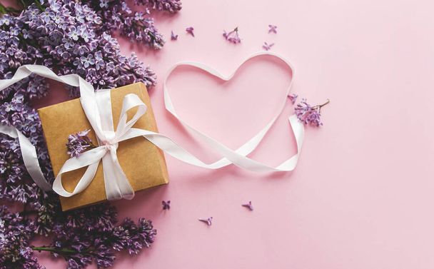 Lilac flowers, gift box and heart ribbon on pink paper, flat lay with copy space. Stylish floral greeting card. Happy valentines day and mothers day concept. Purple lilac bouquet and craft present - Photo, Image