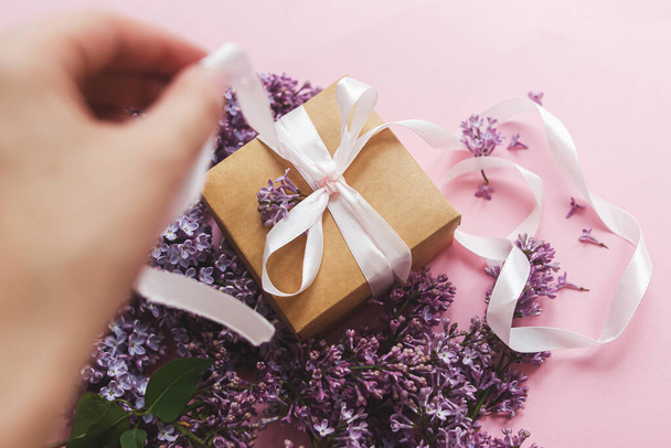 Hand opening gift box with ribbon and lilac flowers on pink paper, view above. Happy mothers day and valentine's day concept. Purple lilac flowers bouquet with craft present box - Photo, Image