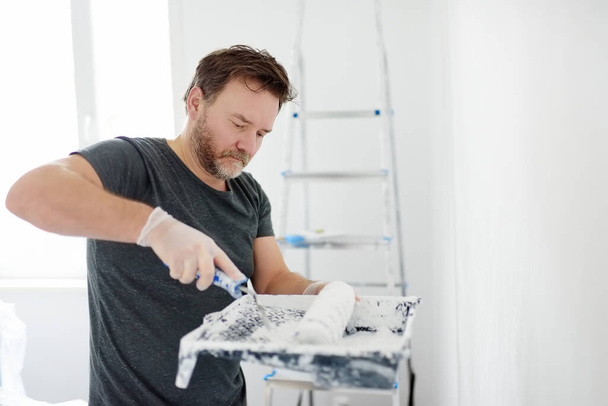 Portrait of a handsome mature man making repairs in the apartment. The person paints the wall white with roller. Do it yourself. DIY. Repair with their hands. - Fotoğraf, Görsel