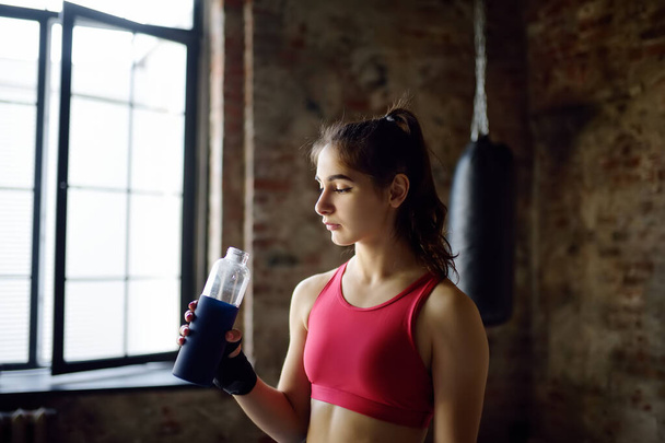 Portrait of young beautiful athlete in a boxing gym. A boxer girl drinks beverage from a bottle during a workout. In the process of training need to drink a lot water. Healthy lifestyle - Foto, imagen