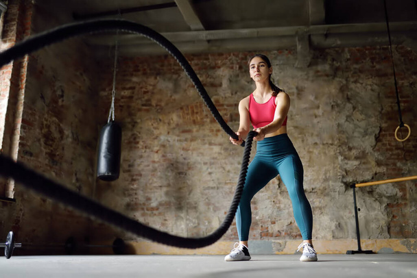 Young woman exercising with battle ropes at the gym. Athletic girl wearing red t-shirt training with battle ropes indoors. Healthy lifestyle - Foto, imagen