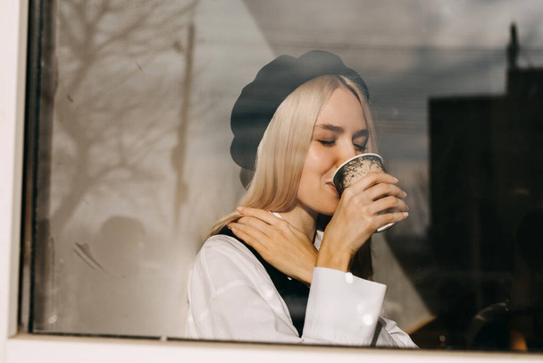 Young stylish woman at a cafe, drinking coffee, sitting by the window in sun light. - Photo, Image