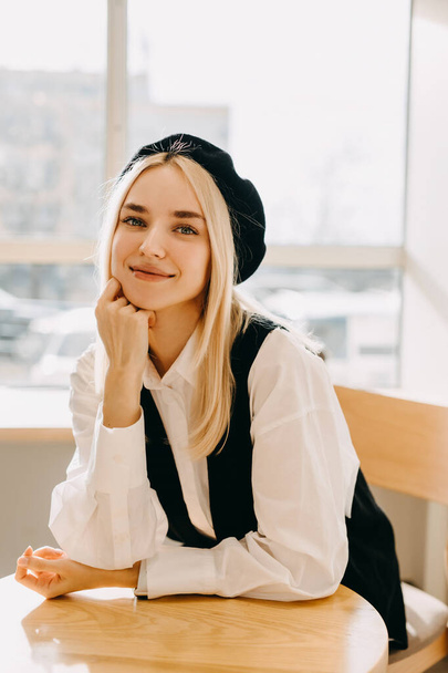 Portrait of a young stylish woman sitting at a cafe table, looking at camera and smiling.  - Photo, Image
