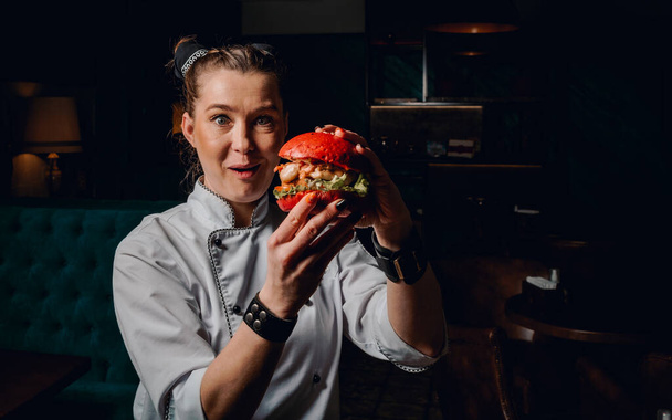 young woman chef with burger in hands on dark background in cafe - Φωτογραφία, εικόνα